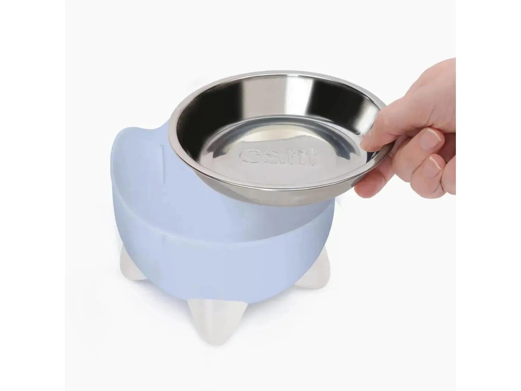 Hånd som plasserer en Catit PIXI vannfontene i lysblå plast med en stål matskål.