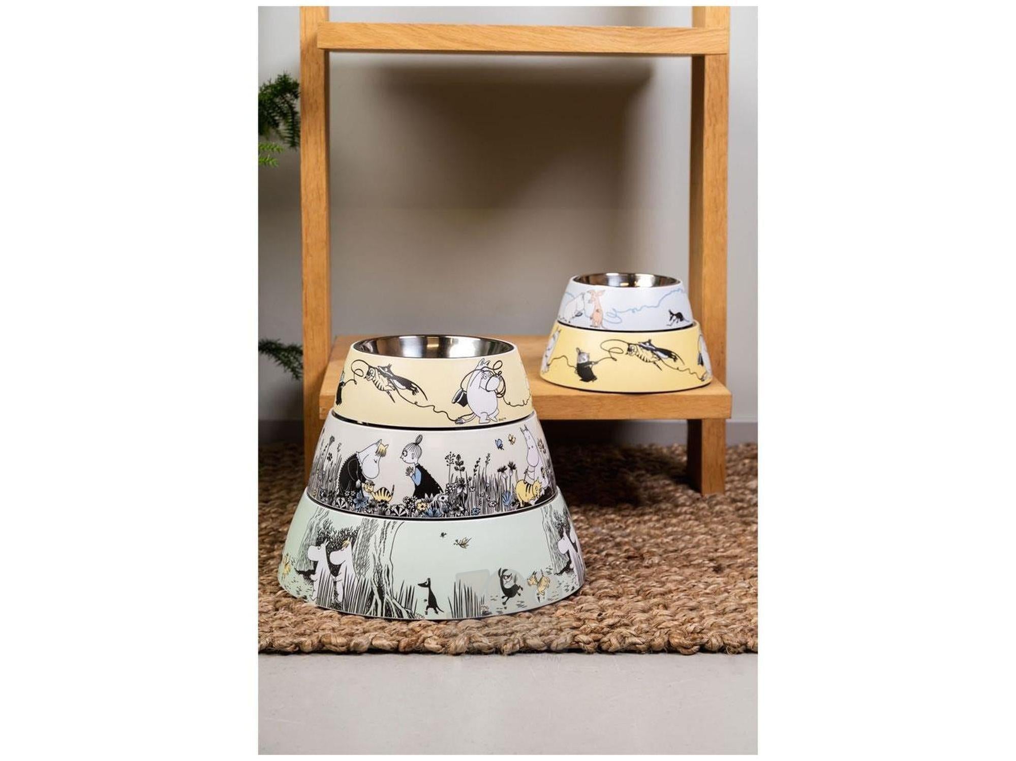 Tre Mummi Pets matskåler i blå, gul og grønn med Mummi-motiv, stablet på et trehyllesystem.
