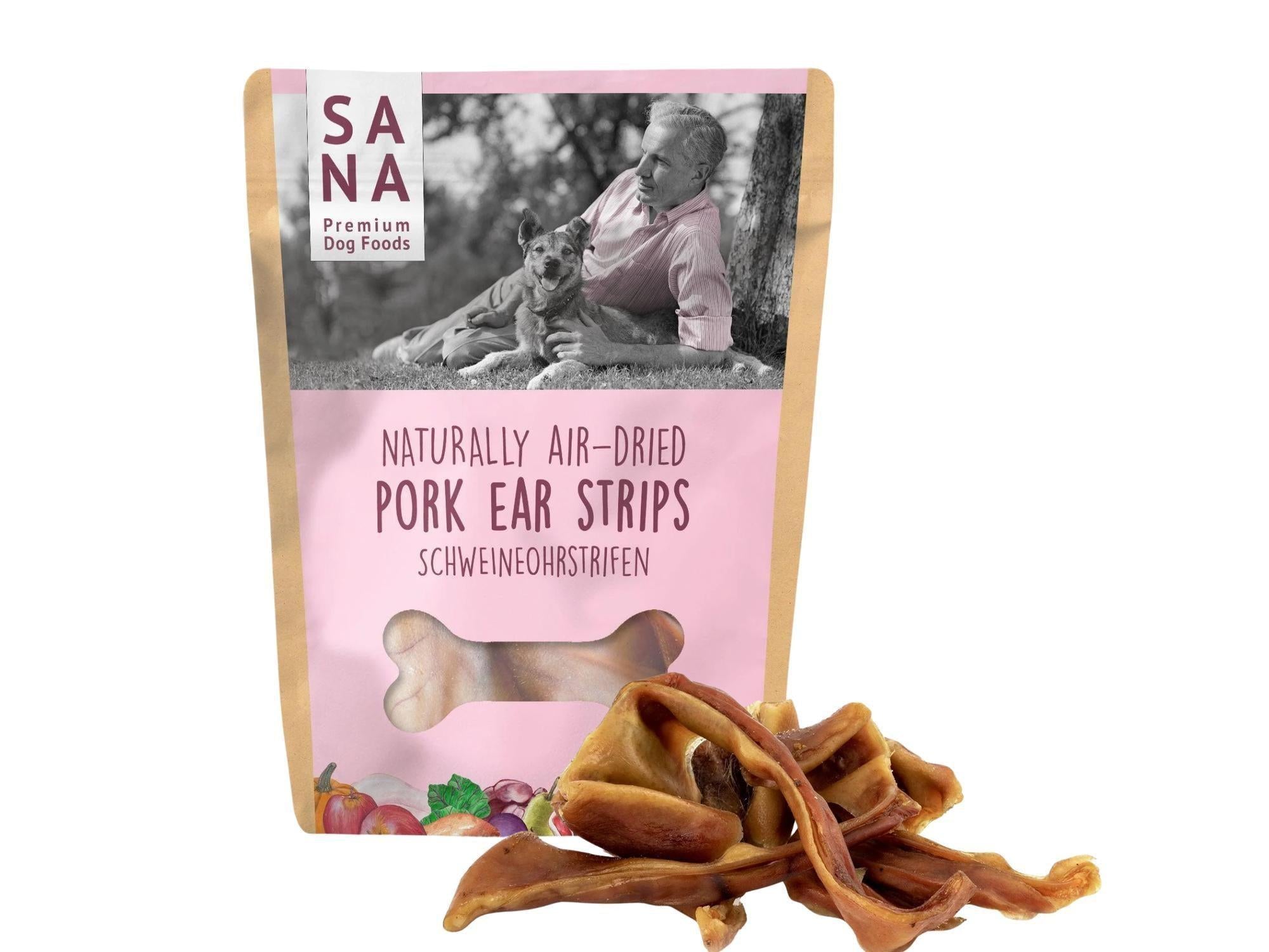 Sana Premium Dog Foods pose med naturlig lufttørkede griseørestrimler og en haug med godbiter.