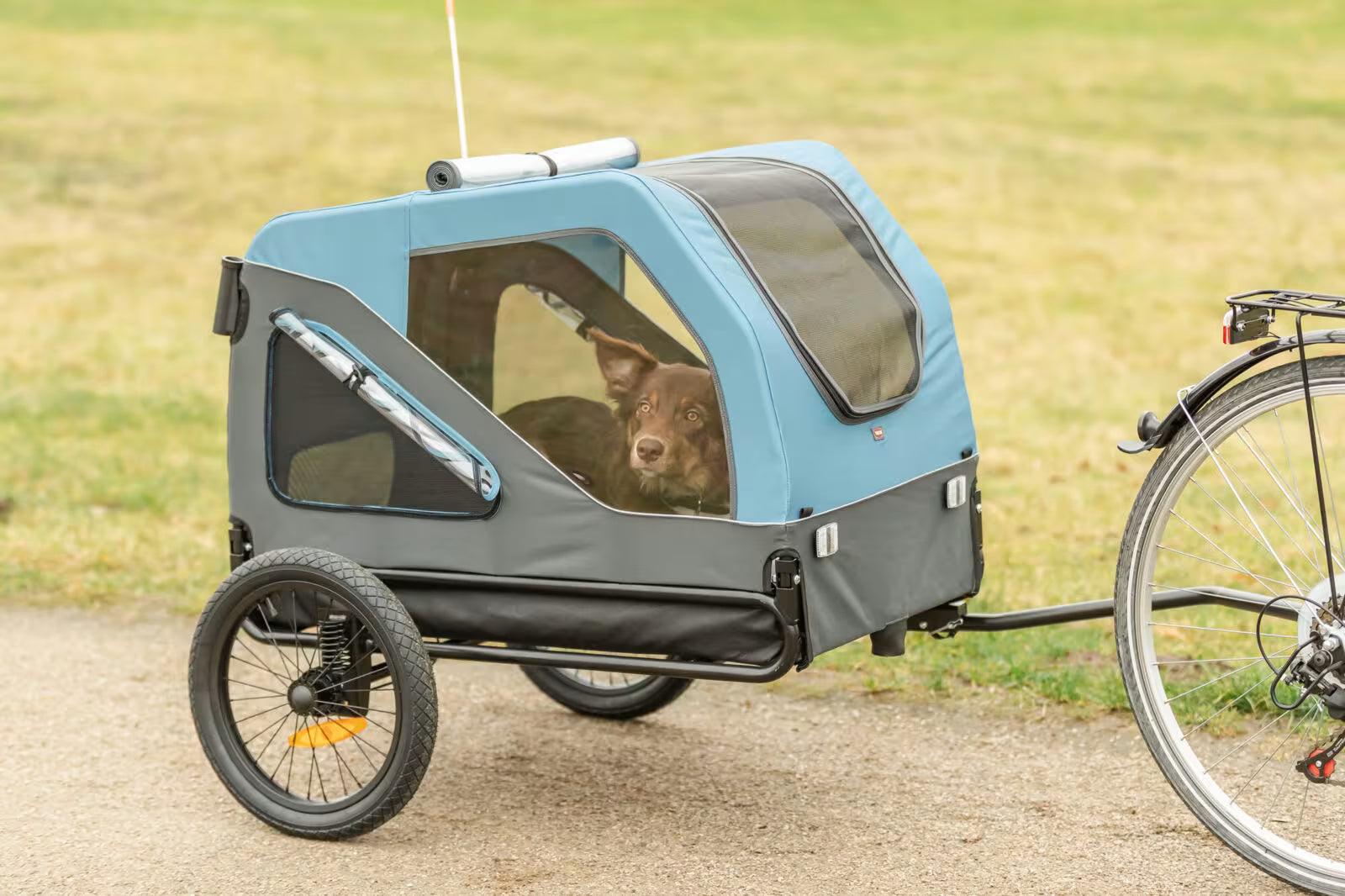 Blå og grå Trixie sykkelvogn for hund med nettingvinduer, festet til en sykkel på en grusvei.